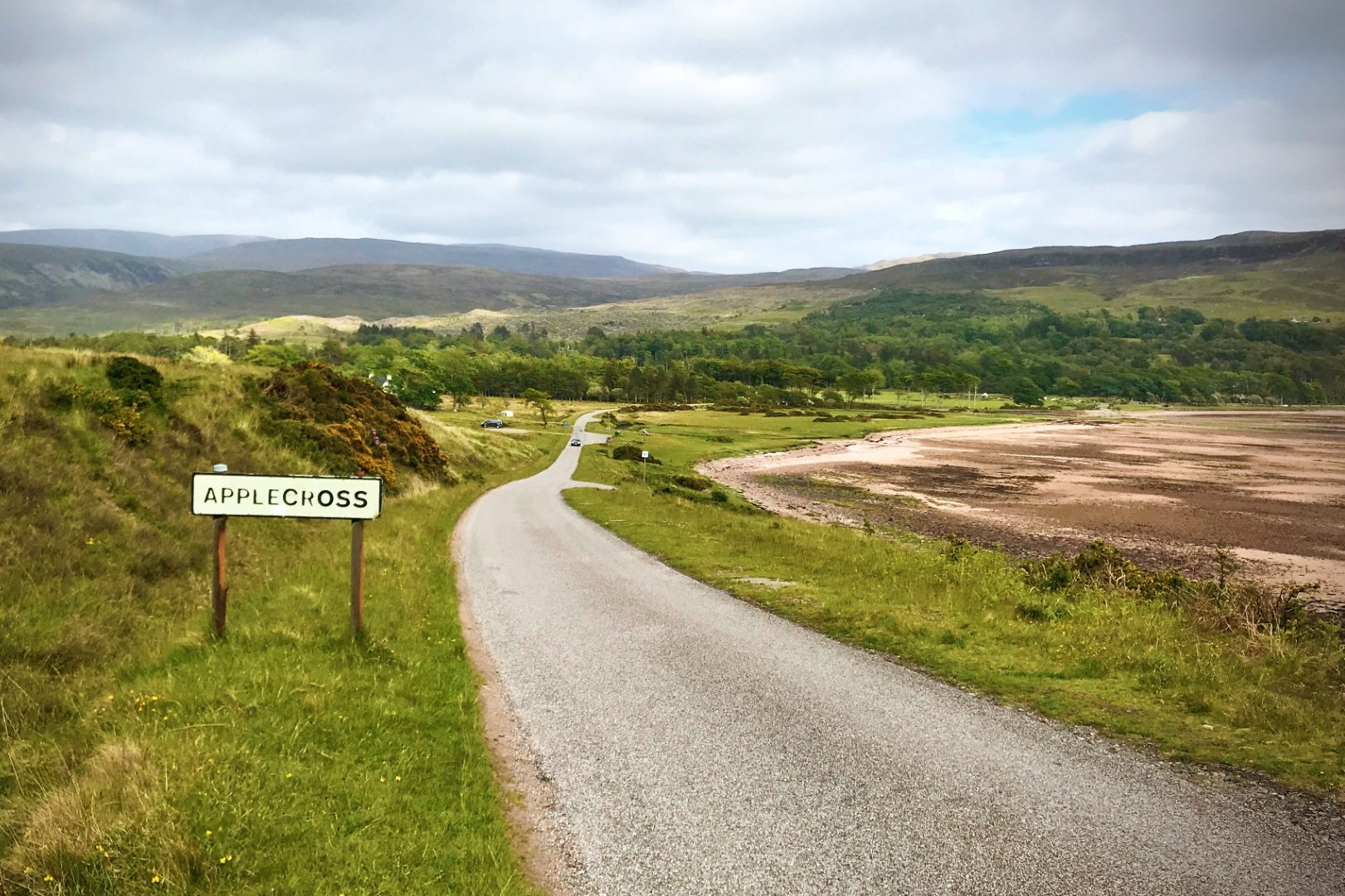 Kurvige Straße durch grüne Landschaft Schottlands, mit einem Schild und Bergen im Hintergrund.