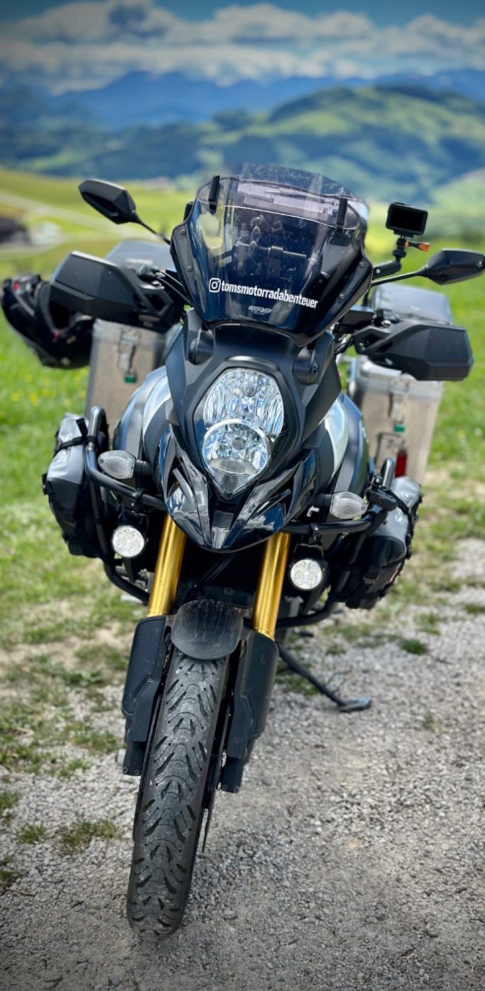 Schwarze Suzuki V-Strom mit Gepäcktaschen, auf einem Feld mit Bergen im Hintergrund.