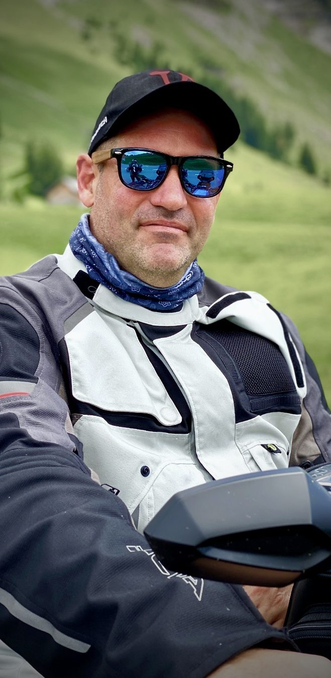 Mann in Motorradjacke und Sonnenbrille, sitzt auf seinem Motorrad inmitten einer grünen Berglandschaft.