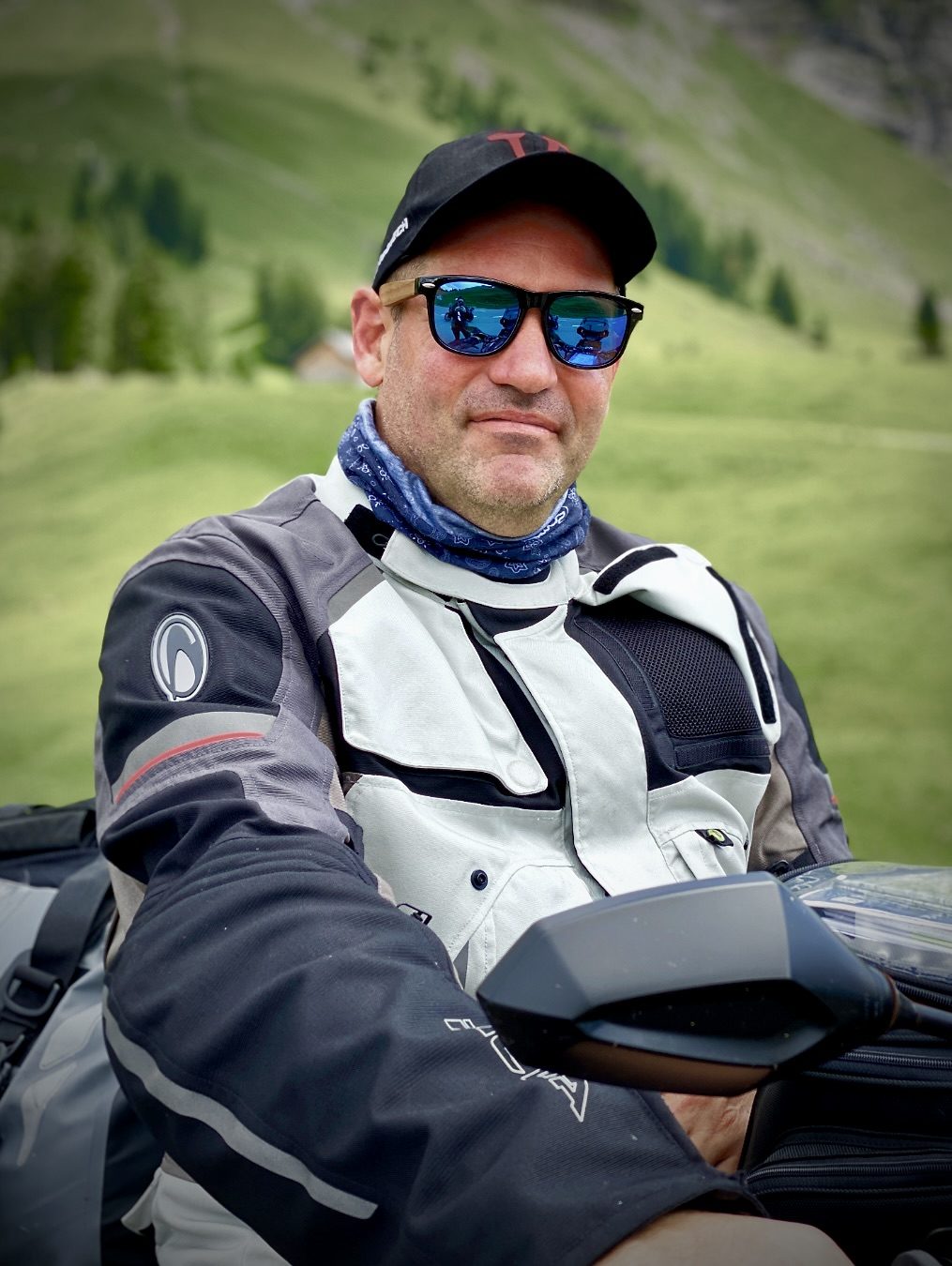Mann mit Sonnenbrille und Motorradjacke, sitzt im Freien vor grünen Hügeln.