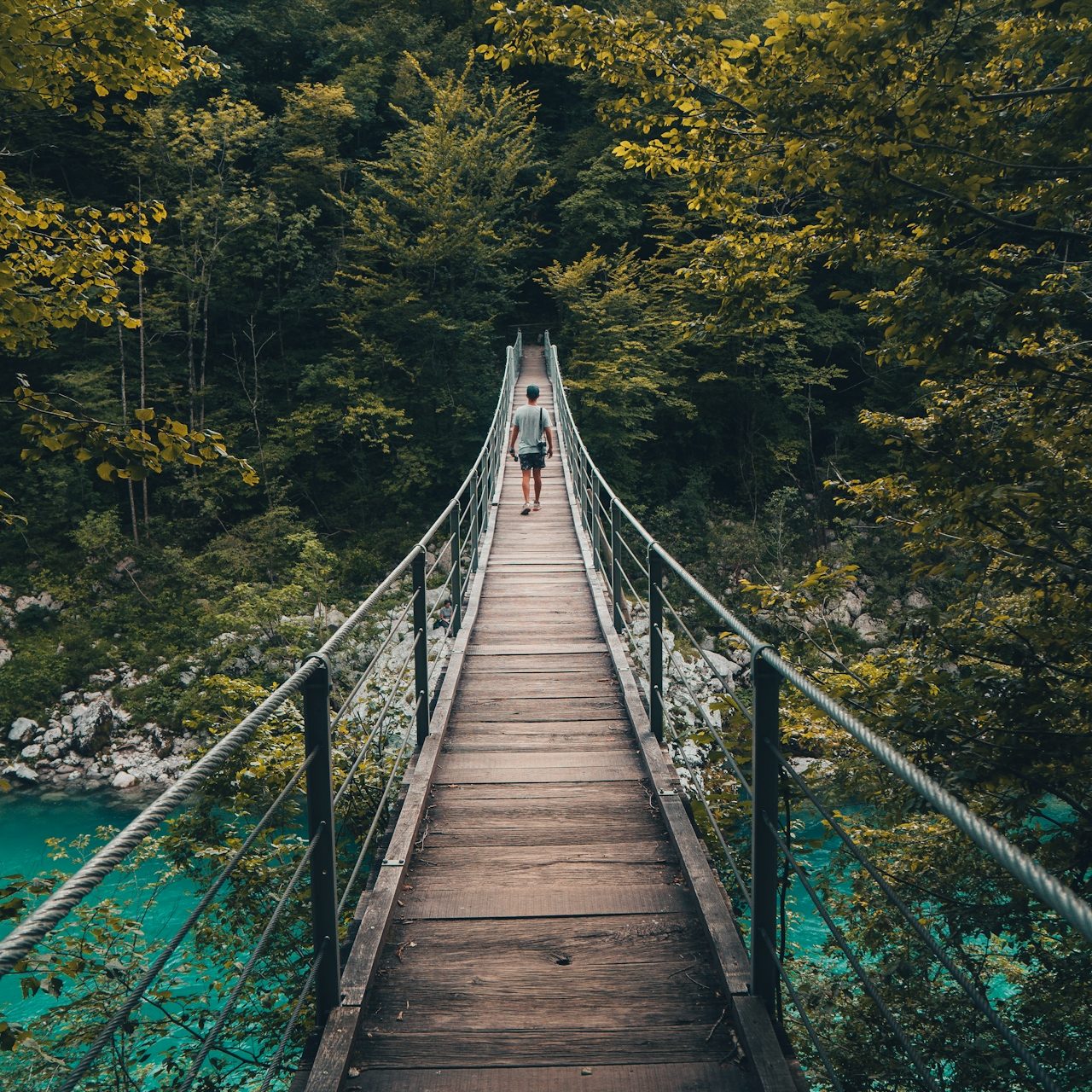 Eine Holzbrücke über einen türkisfarbenen Fluss, umgeben von grünen Bergen und Bäumen.
