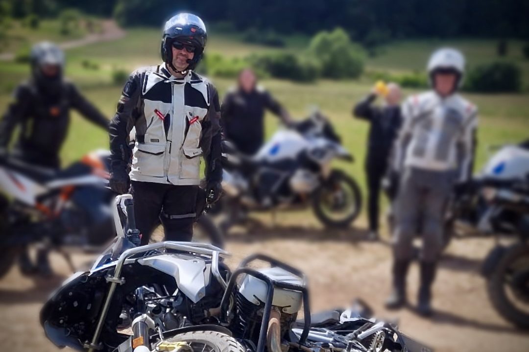 Gruppe von Motorradfahrern auf einer grünen Wiese, einige stehen hinter einem umgefallenen Motorrad.